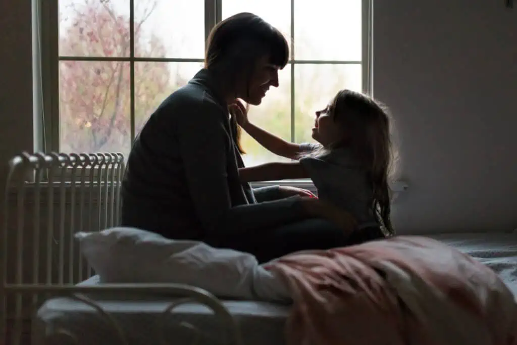 When you show your child you love them, that has a lifelong positive impact. Pictured: A mom and her daughter sitting face-to-face while sitting on a bed, the child reaching out lovingly to the mother.